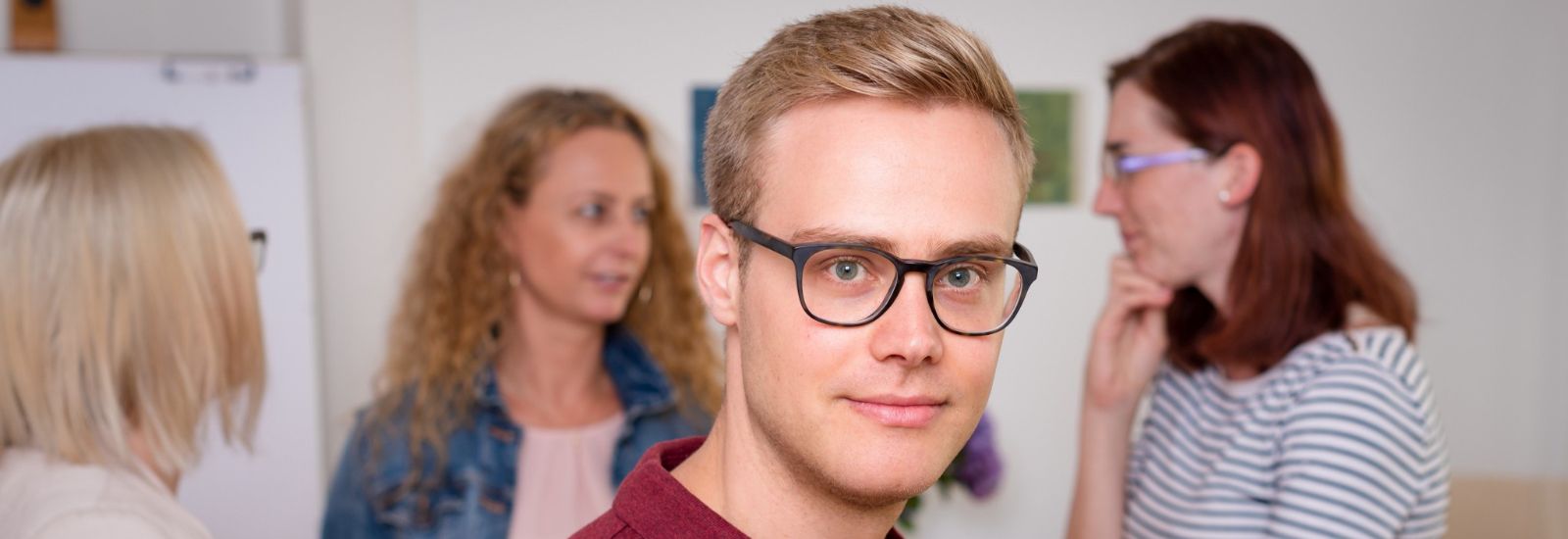 Ein Mann mit Brille blickt in die Kamera, drei Frauen unterhalten sich im Hintergrund.