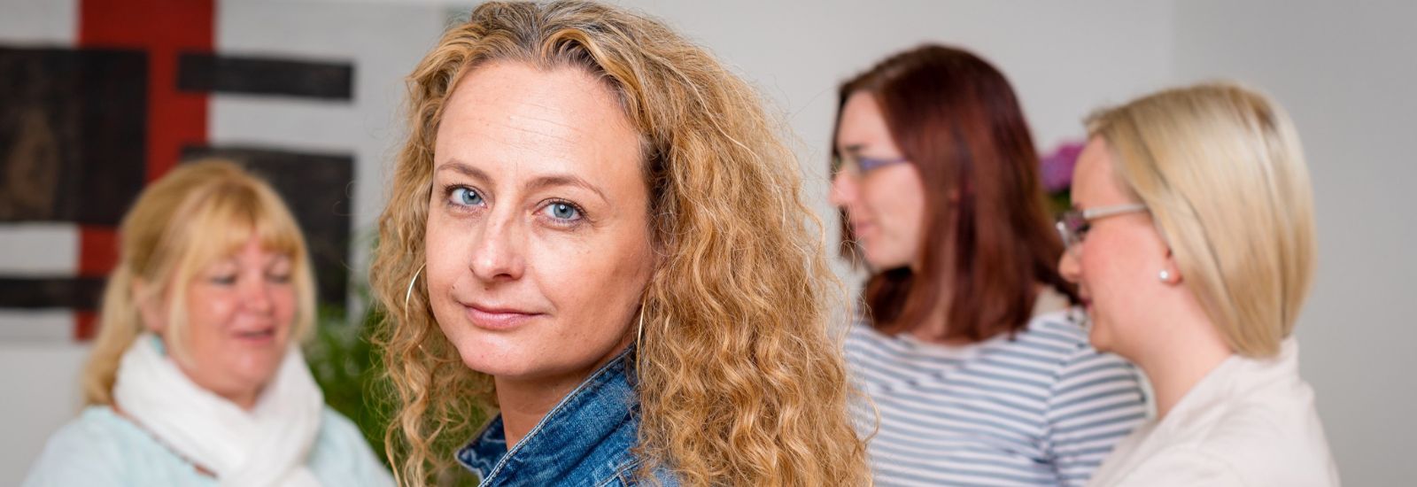 Eine Frau mit lockigem Haar blickt in die Kamera, drei weitere Frauen unterhalten sich im Hintergrund.
