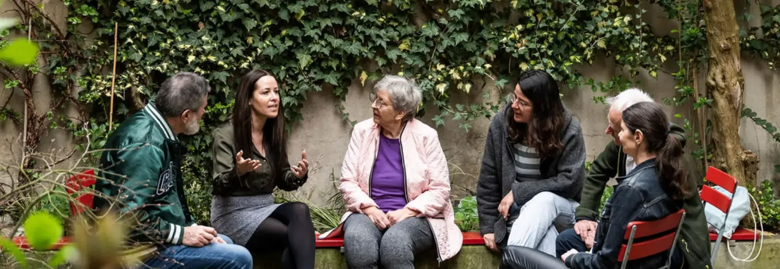 Eine Gruppe von sechs Personen unterschiedlichen Alters sitzt im Kreis in einem grünen Garten und hört aufmerksam einer Frau zu, die mit ausdrucksstarken Gesten spricht.